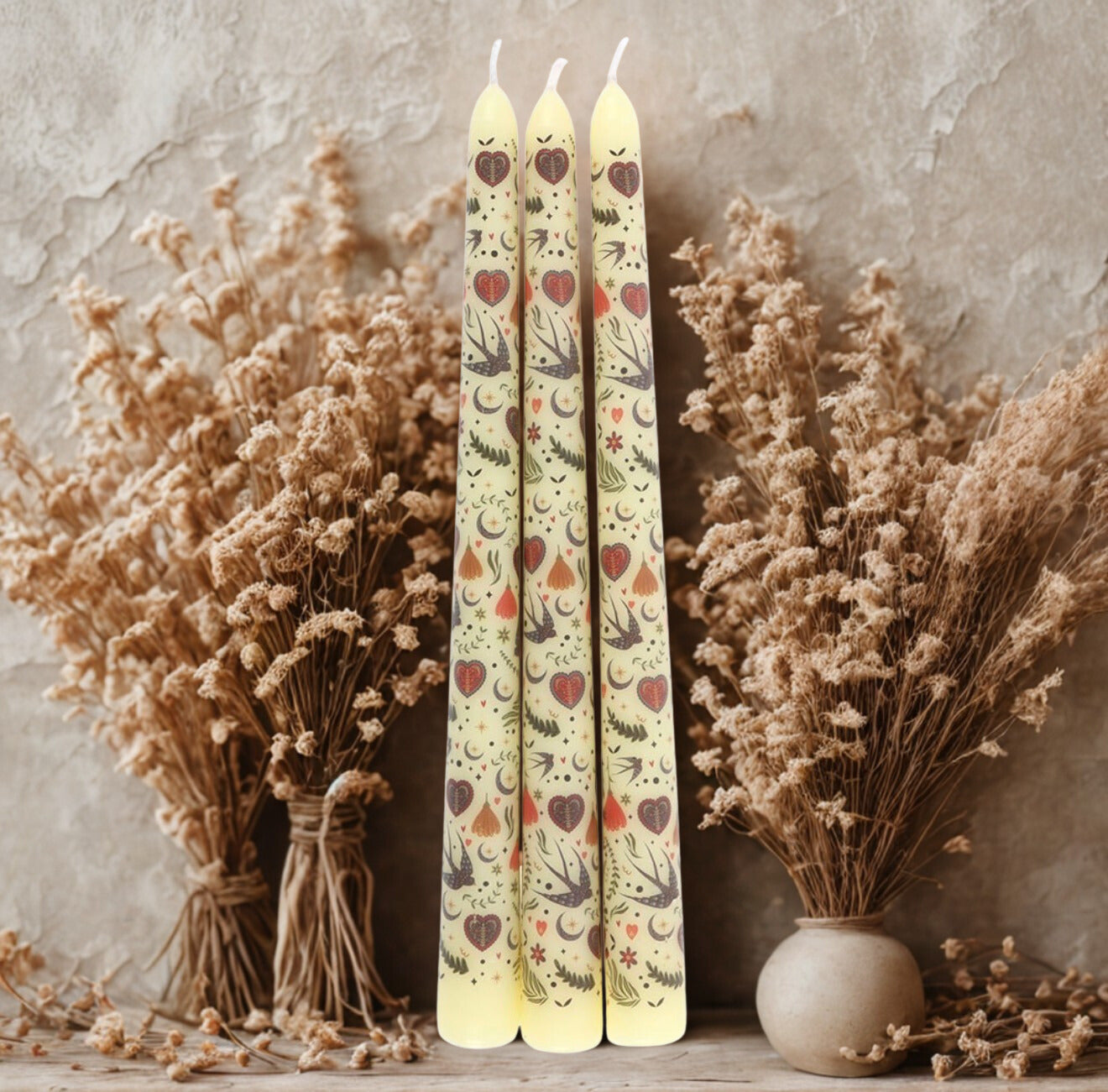 Sfeervolle set van drie dinerkaarsen met een gedetailleerde Folk-stijl bedrukking, liggend op een zachte beige ondergrond van droogbloemen voor een natuurlijke uitstraling.