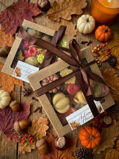 ⁠Gestapelde Autumn Harvest wax melts cadeaudoosjes met bruin lint op een sfeervolle houten herfstavond achtergrond met bladeren, kastanjes en kaarslicht⁠