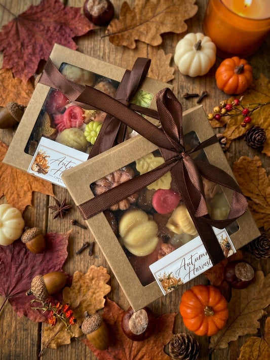 ⁠Gestapelde Autumn Harvest wax melts cadeaudoosjes met bruin lint op een sfeervolle houten herfstavond achtergrond met bladeren, kastanjes en kaarslicht⁠