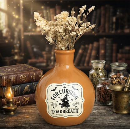 Een mysterieus tafereel voor Halloween, met centraal een oranje brouwselfles met het etiket 'FOR CURSING TOADBREATH'. De fles is gevuld met een bosje gedroogde bloemen en heeft een koperen houder met een zwarte kaars erin, wat zorgt voor een dubbele functie als vaasje en kandelaar. Het staat op een houten oppervlak, geflankeerd door vintage boeken en een olielamp aan de linkerkant, en glazen potten met ingrediënten en een vijzel aan de rechterkant. De achtergrond is een donkere bibliotheek.