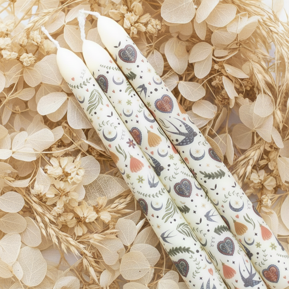 Drie decoratieve dinerkaarsen met bloemen-, hartjes- en vogelmotief liggend op een rustige achtergrond van gedroogde beige bladeren en takken.