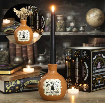 Een sfeervolle close-up foto van een oranje keramische heksenfles, perfect voor Halloween-decoratie. De fles is multifunctioneel en doet dienst als vaasje met gedroogde bloemen én als kandelaar, met een zwarte kaars in een koperen houder die uit de hals steekt. Het crèmekleurige etiket draagt de tekst "FOR CURSING TOADBREATH" met een illustratie van een kikker met een heksenhoed en vleermuizen. Het object staat op een rustieke houten tafel, omringd door oude spreukenboeken, een brandende olielamp en glazen 
