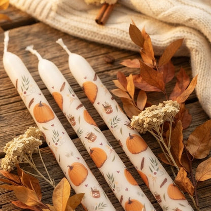Een set van drie tapstoelopende kaarsen met een herfstthema, versierd met illustraties van pompoenen, mokken en bladeren, geplaatst op een rustieke houten tafel met herfstdecoraties.