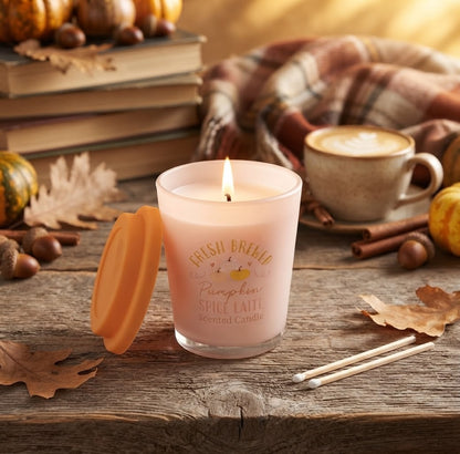 Een close-up van een gezellige herfsttafelsetting. Een roze 'Fresh Brewed Pumpkin Spice Latte' geurkaars straalt warm licht uit, met het oranje deksel er bovenop geplaatst. Een rokend kopje koffie met kaneel staat ernaast. De scène is gevuld met het zachte zonlicht van een herfstochtend, die de talloze mini-pompoenen, kaneelstokjes, steranijs en droogbloemen op de achtergrond verlicht.