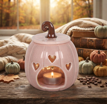 Een roze keramische pompoen-vormige wasbrander met hartvormige uitsparingen, brandend theelichtje binnenin en bruine steel. Geplaatst op een rustieke houten tafel met herfstdecoraties.