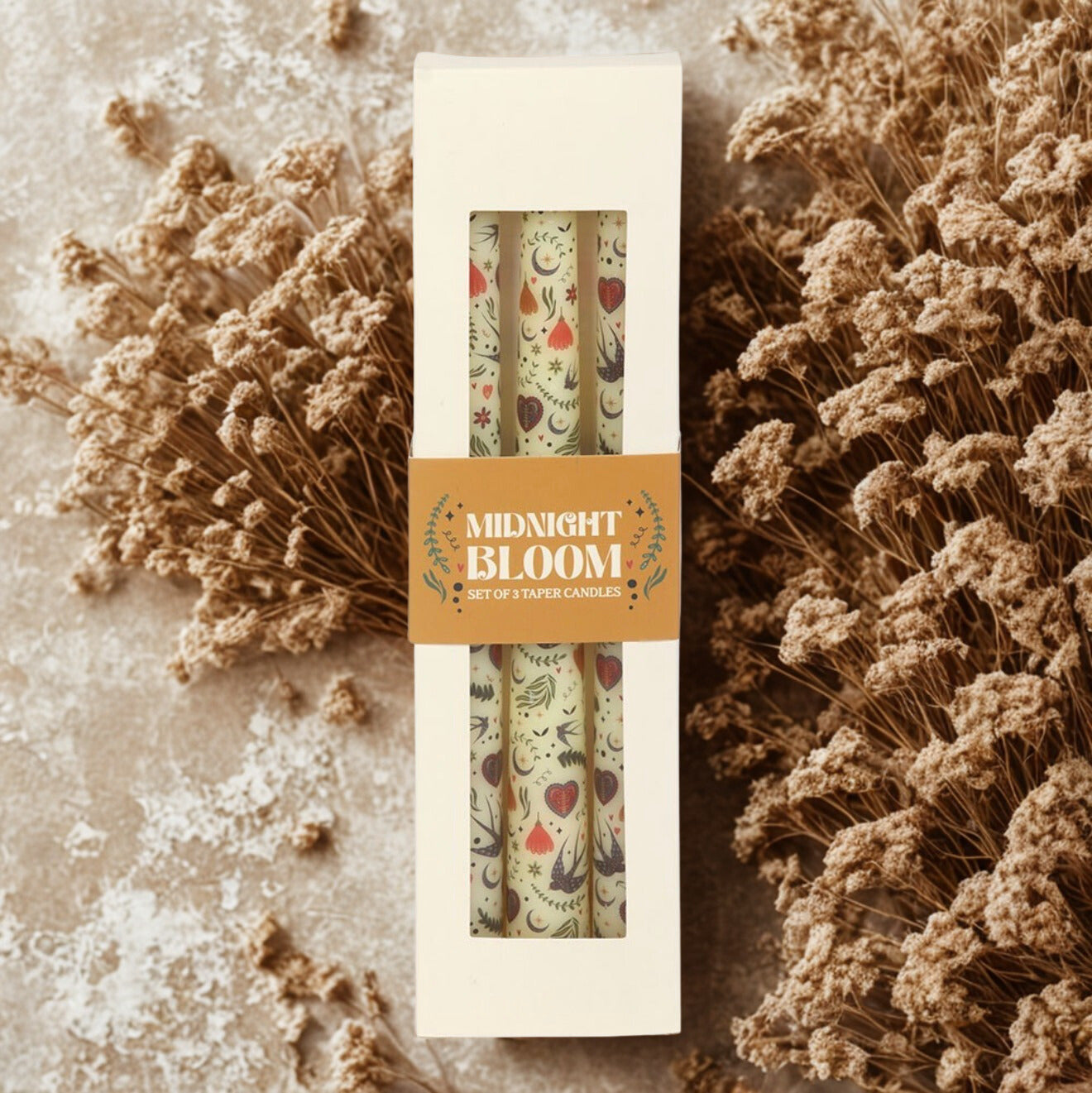 Close-up van een set van drie ongeparfumeerde dinerkaarsen met een folkloristische print van bloemen en maantjes, gepresenteerd op een natuurlijke, beige achtergrond.